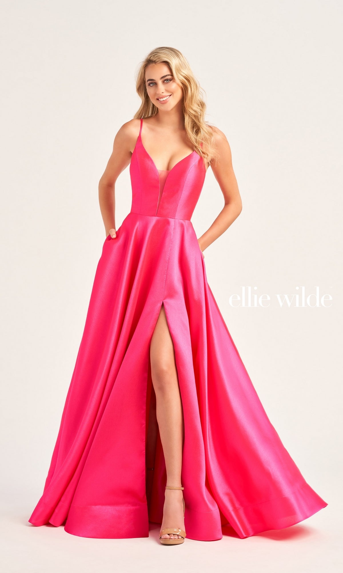 A woman poses with her hands in the pockets of the Ellie Wilde Long A-Line Satin Prom Dress EW35232, a bright pink sleeveless gown featuring a thigh-high slit, set against a plain background.